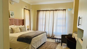 A hotel room with a bed, a desk, a chair, a small table, and curtains. - Mmaset Houses 3-bedrooms (Gaborone)