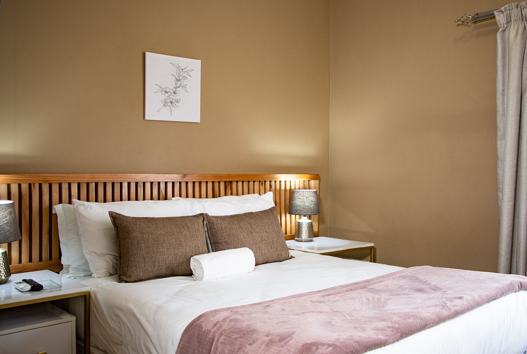 Una cama bien hecha con ropa de cama blanca y marrón, flanqueada por dos mesitas de noche con lámparas, un cabecero de madera y una obra de arte colgada en la pared.