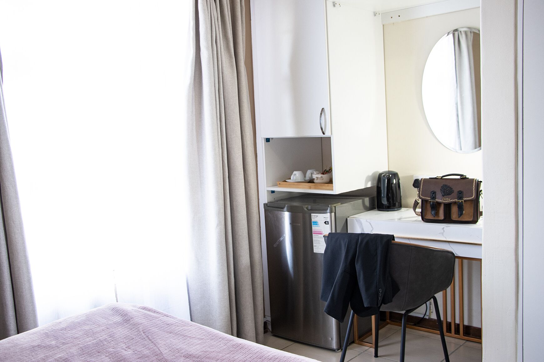 Habitación de hotel con cama, escritorio con cafetera, maleta y refrigerador.