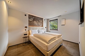 A modern bedroom with a large bed, a small side table, and a view of greenery through a window.