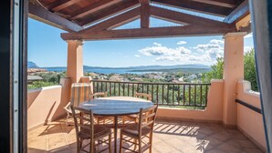 A covered outdoor patio with a wooden table and chairs, a view of a coastal landscape, and a railing.