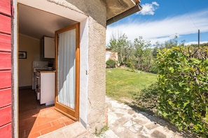 A small kitchen with cabinets and a refrigerator, a wooden door leading to a patio, and a garden with grass and shrubs. - Farmhouse 'Podere Del Pereto Bio- 202' with Shared Pool, Private Garden and Wi-Fi (Rapolano Terme)