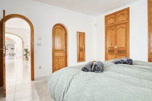 A bedroom with a bed, two wooden doors, a hallway, and a kitchen area.