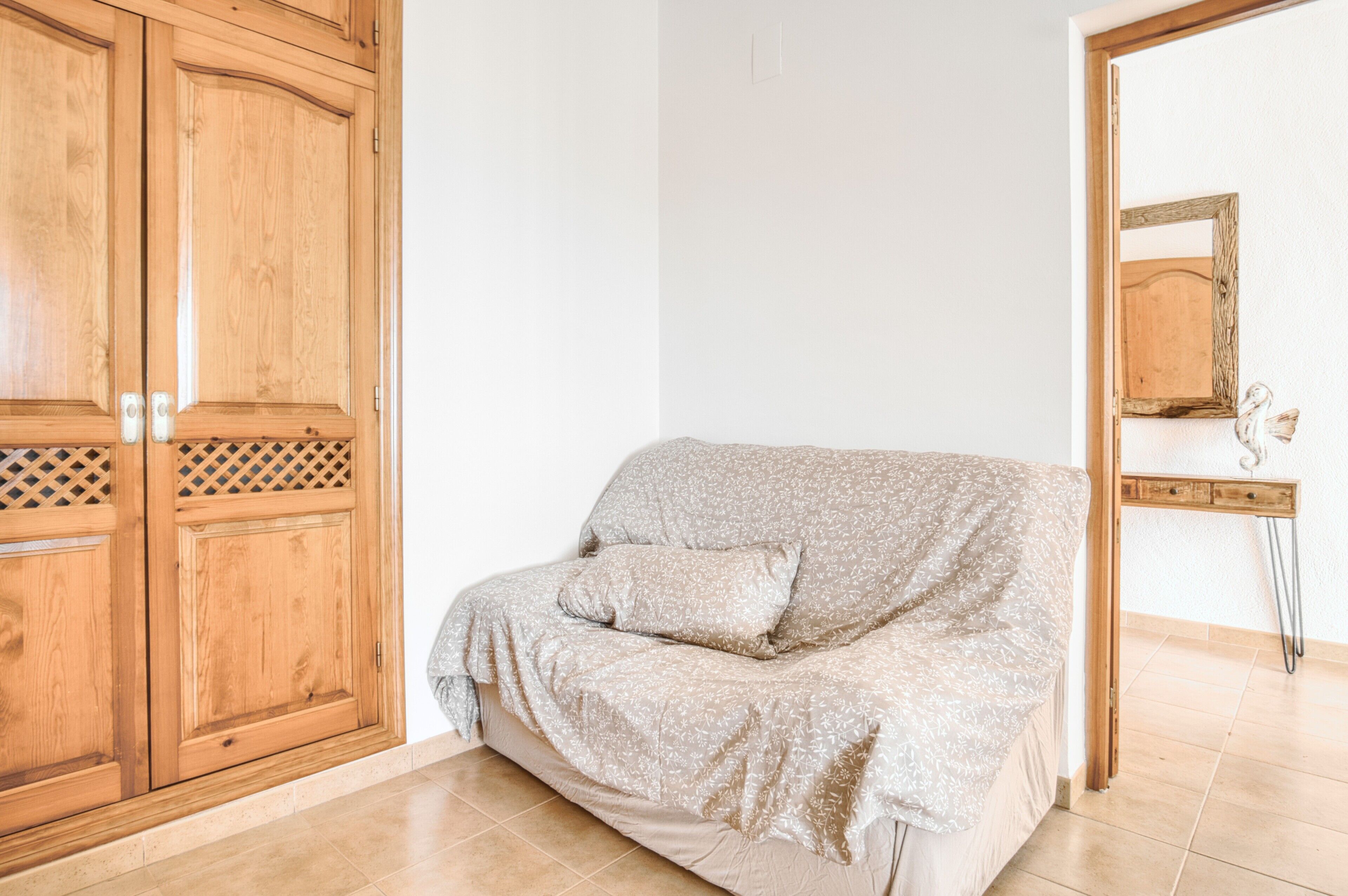 A room with a wooden wardrobe, a sofa with a patterned cover, and a mirror on the wall.