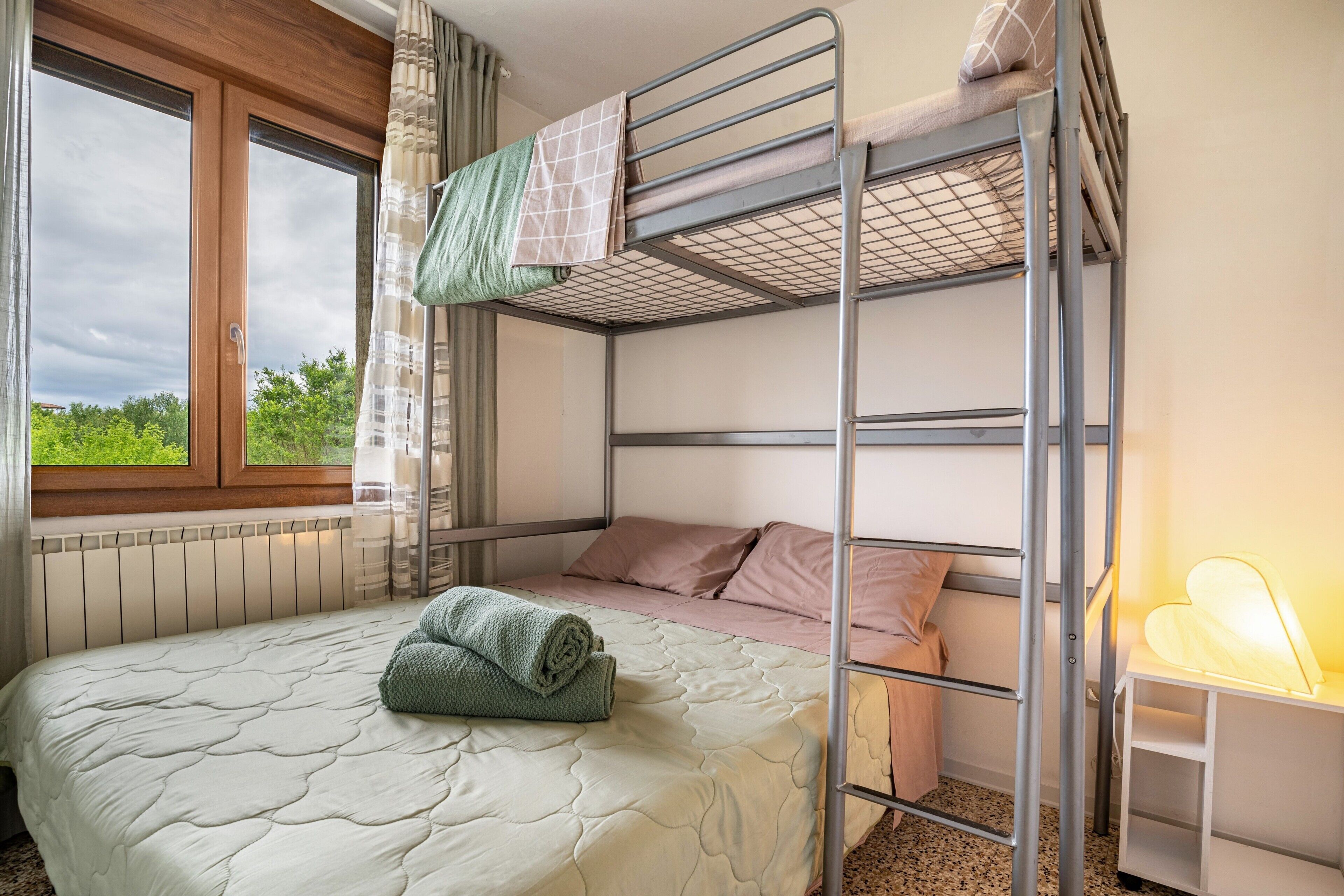 A bunk bed room with a window, a bedside table with a lamp, and a rolled-up blanket on the lower bed.