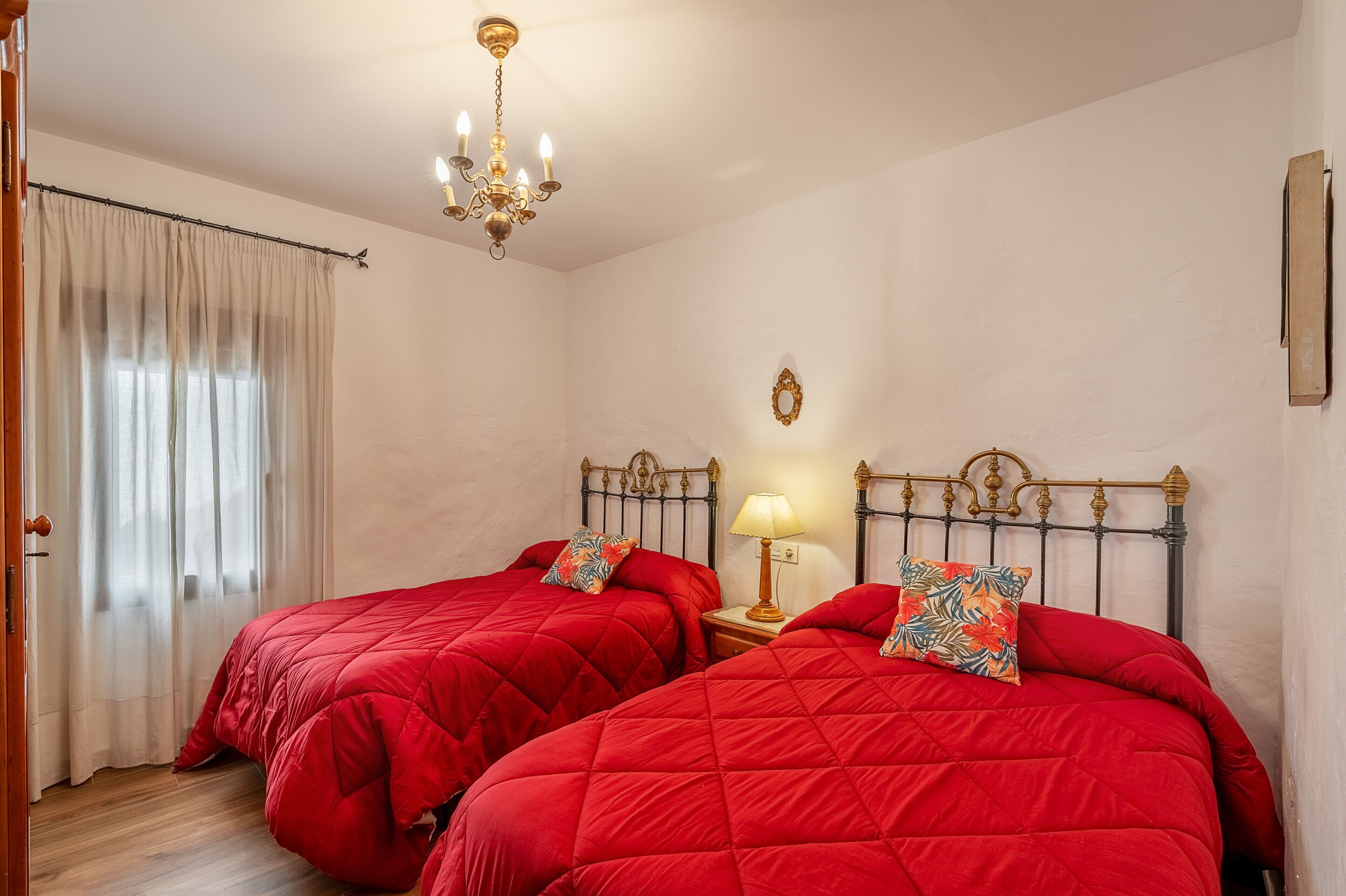Quarto com duas camas, um lustre, mesa de cabeceira com abajur e janela com cortinas.