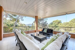 A covered balcony with wicker furniture, a glass coffee table, and a view of trees and hills. - Holiday Home 'La Brisa' with Sea View, Wi-Fi and Air Conditioning (Llafranc)