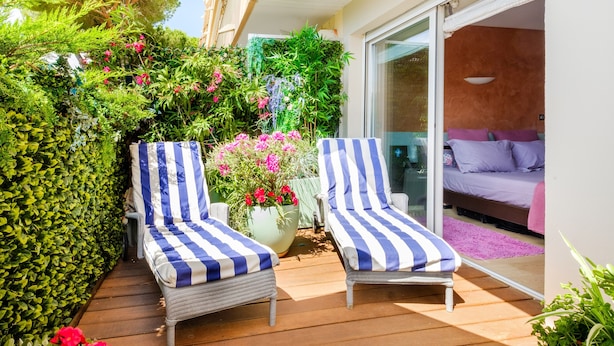 A balcony with two striped lounge chairs, a potted plant with pink flowers, and a sliding glass door leading to a bedroom with a purple bedspread. - Apartment 'Des Vacances De Rêve À Mandelieu' with Shared Pool and Air Conditioning (Mandelieu-la-Napoule)