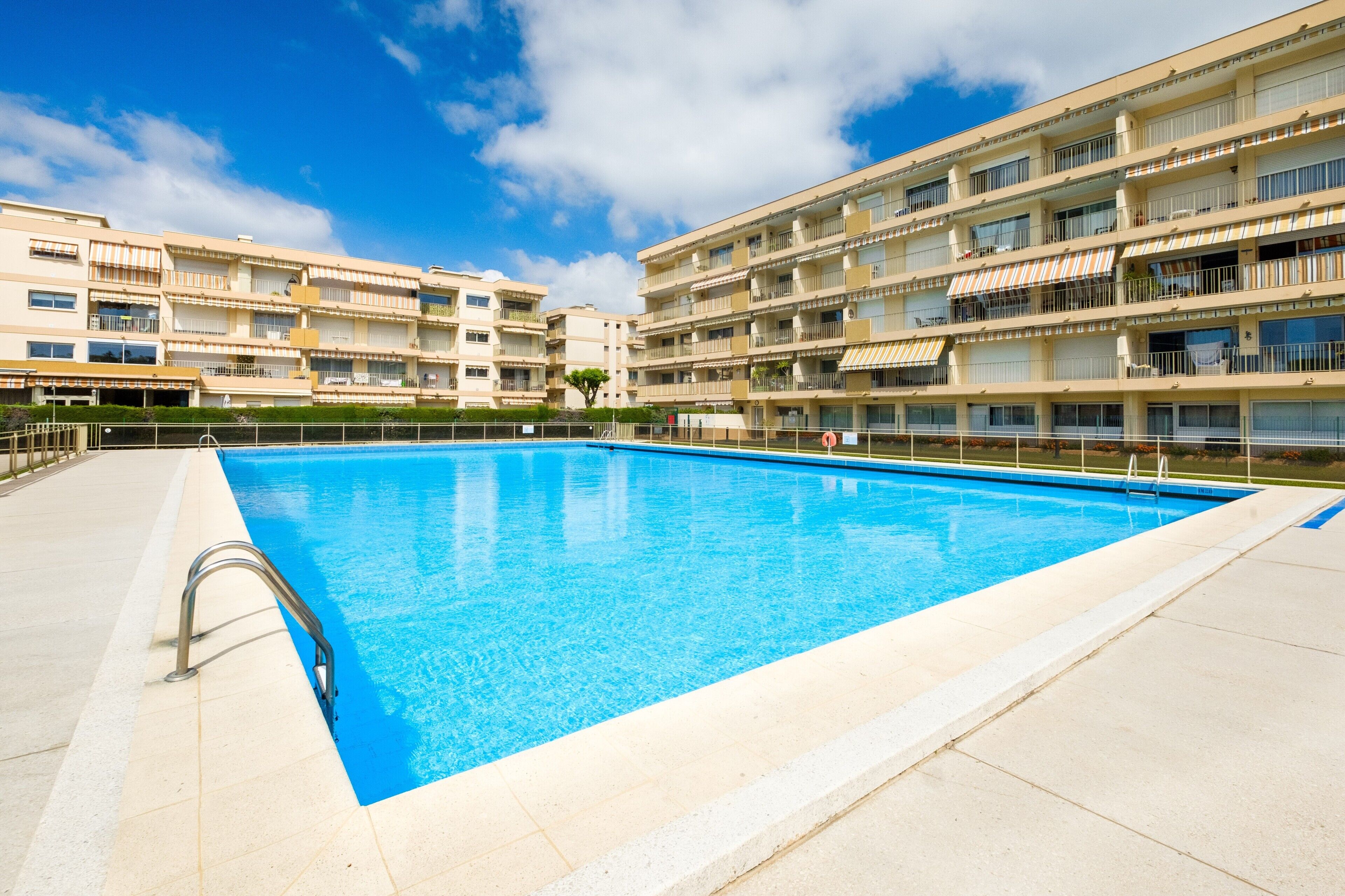 Une piscine équipée d’un escalier en métal, entourée d’une terrasse carrelée et jouxtant un immeuble à plusieurs étages doté de balcons.