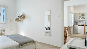 A bedroom with a bed, bedside table, and a mirror. There is a small desk and chair in the corner. A kitchenette is visible in the adjacent room.