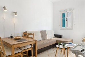 Une pièce au décor minimaliste, avec une table en bois, un canapé, une petite table d’appoint et une fenêtre aux volets bleus.