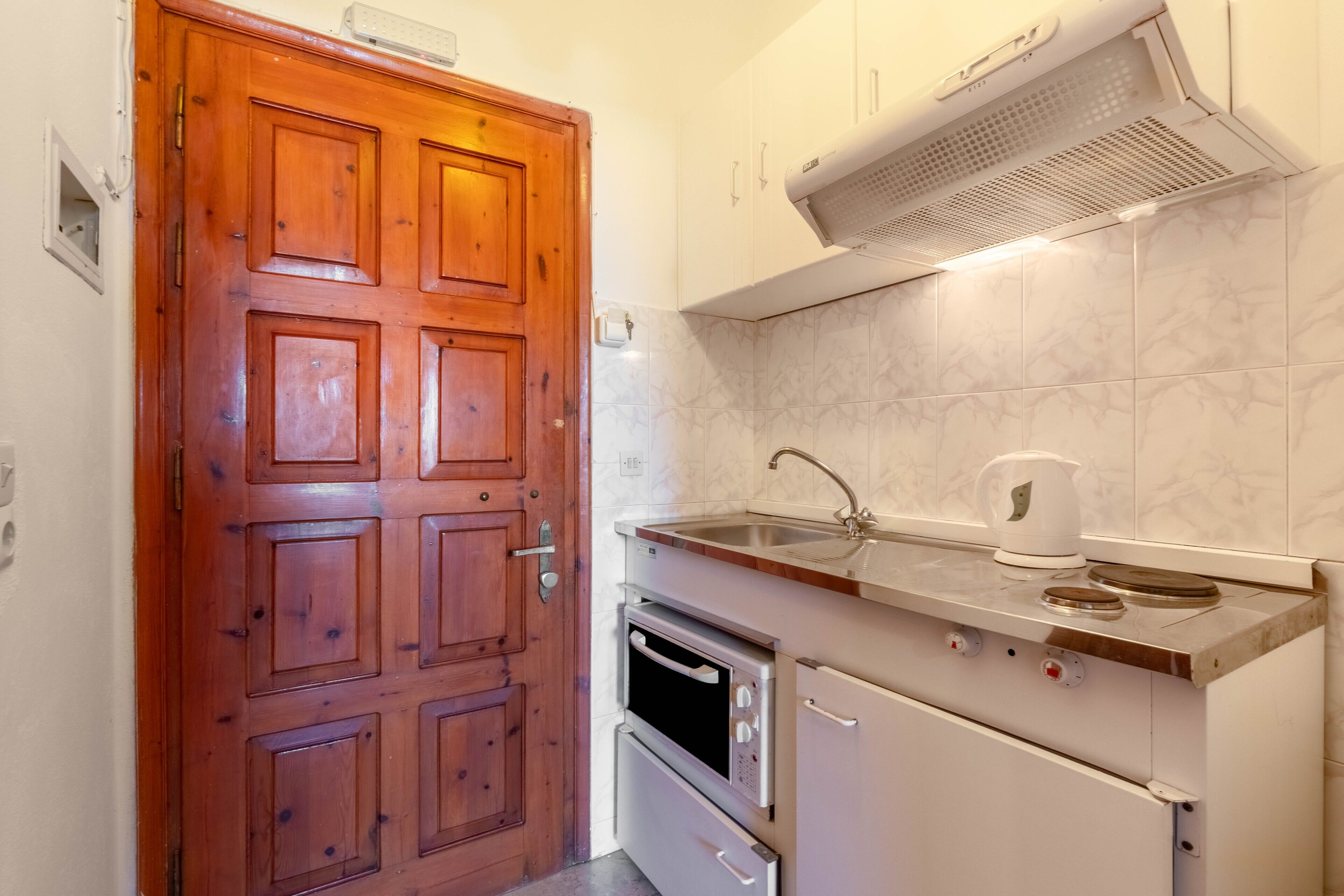 A compact kitchen with a wooden door, a built-in oven, a microwave, a sink, and a kettle.