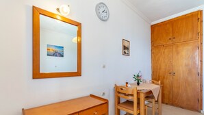 Ein Zimmer mit Schreibtisch aus Holz, einem Stuhl, einem kleinen Tisch mit Tischdecke, einer Uhr und einem Kleiderschrank.