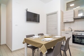 Un comedor con una mesa preparada para cuatro personas, un televisor montado en la pared y una cocina con estufa y horno.