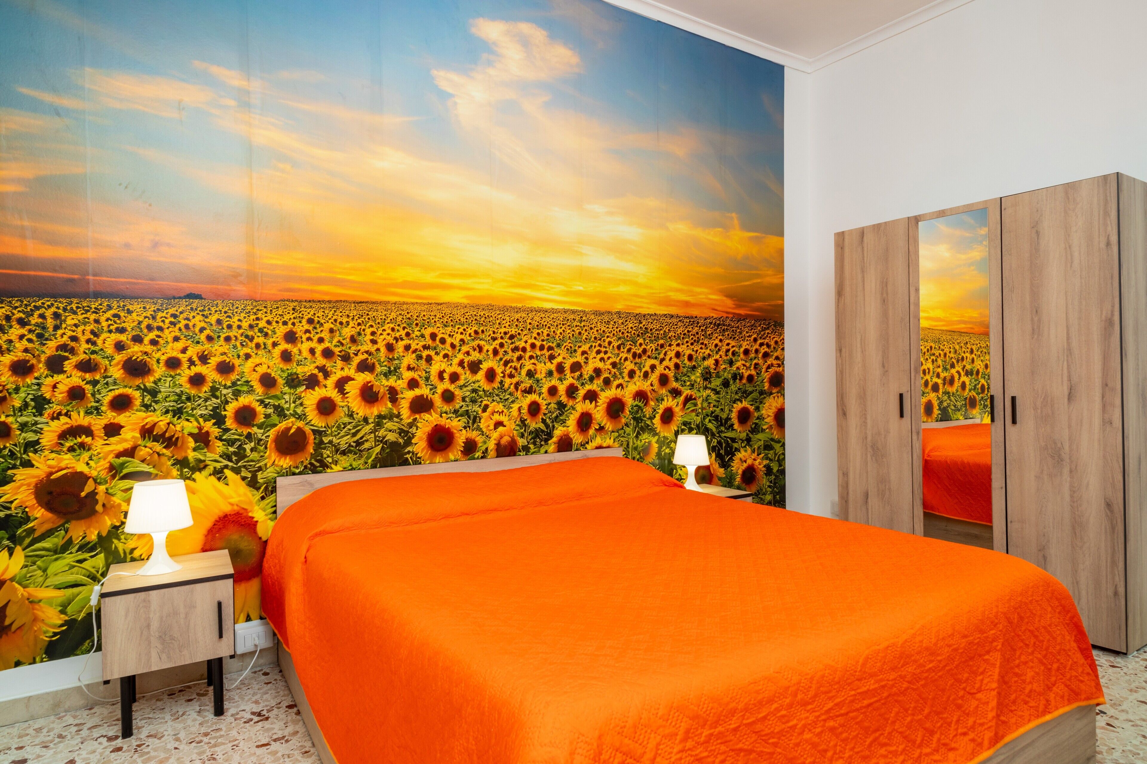 A bedroom with a large mural of a sunflower field, a wooden wardrobe, and a bed with an orange bedspread.