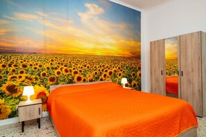 Une chambre avec une grande fresque représentant un champ de tournesols, une armoire en bois et un lit recouvert d’une couverture orange.