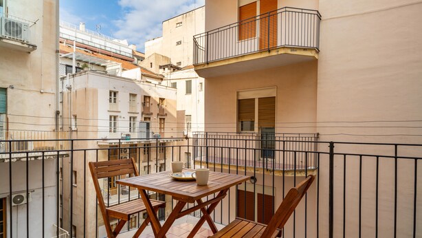 Un balcón con mesa y sillas de madera, vistas a edificios vecinos y baranda de metal.