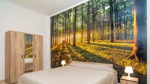 Un dormitorio con un gran mural de un bosque, un armario de madera, una cama con colcha color beige y dos lámparas de noche.