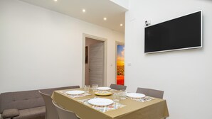 Un comedor con una mesa dispuesta para cuatro, sillas, un sofá y un televisor montado en la pared.