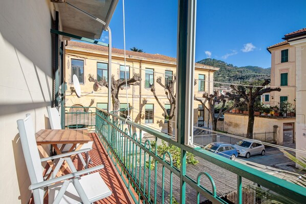 A balcony with a table and chairs, a view of buildings, and a street with parked cars.