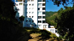 Un immeuble blanc à plusieurs étages, doté de balcons, entouré d’arbres et de collines.