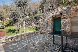 Un patio de piedra con una mesa y una puerta verde con contraventanas, rodeado de olivos y un muro de piedra.