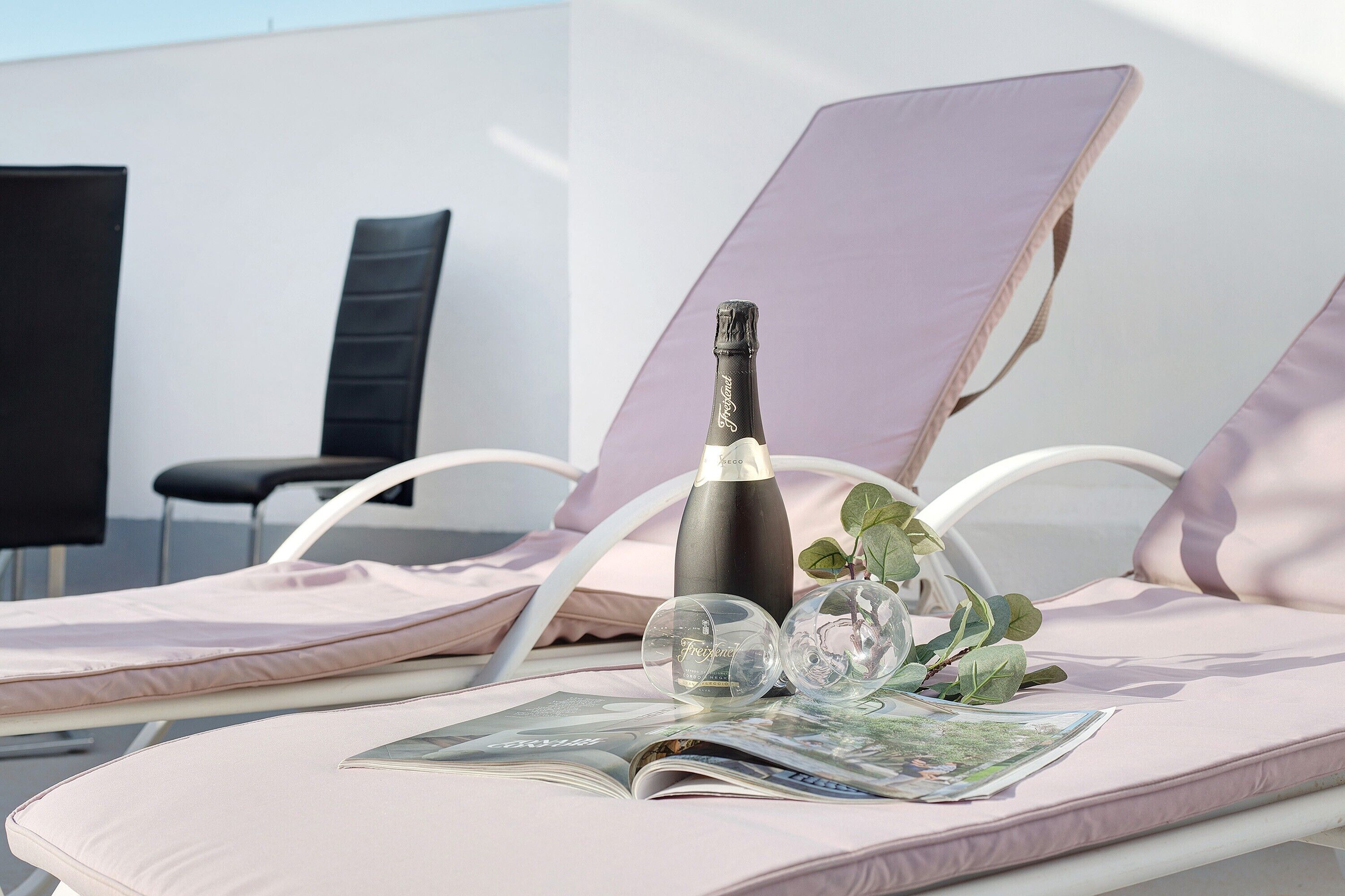 A lounge chair with a magazine, a bottle, and two glasses on a table.