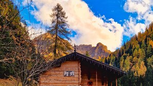 Eine Holzhütte mit einem Balkon, einer Steinsäule links und einer Bergkulisse im Hintergrund.