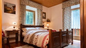 Ein Schlafzimmer mit einem hölzernen Bett, zwei Nachttischen, einem Fenster mit blumigen Vorhängen und einem weißen, flauschigen Teppich.