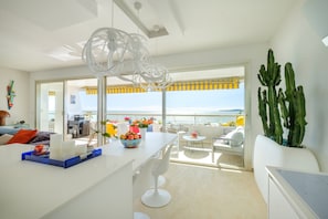 A modern kitchen with a large island, a dining area with white chairs, a cactus plant, and a view of the sea.