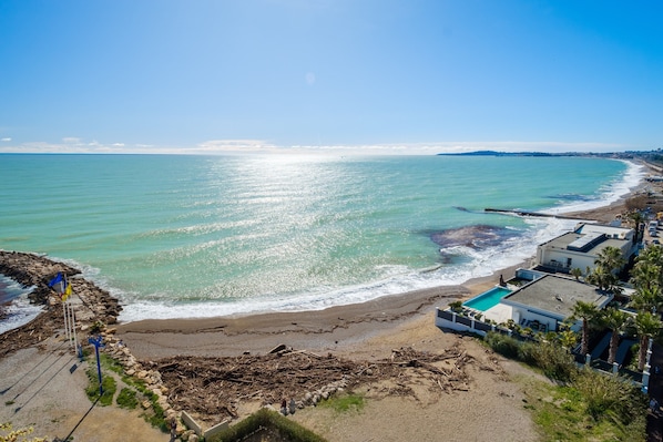 A coastal view with a beach, ocean, and a row of houses with a swimming pool.