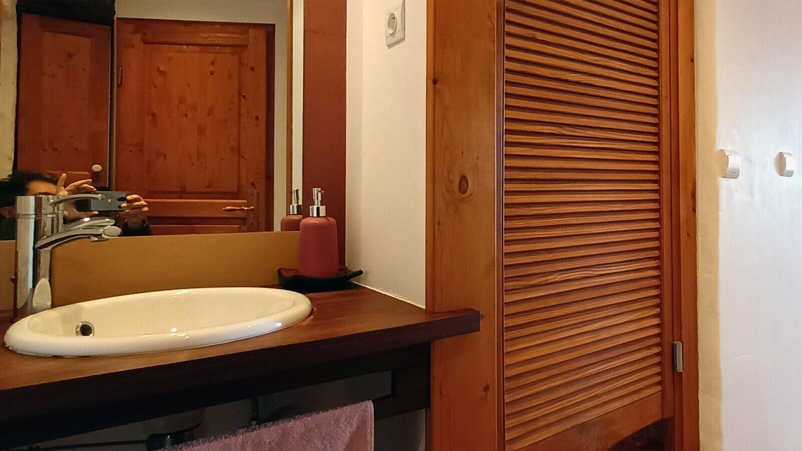 A bathroom with a sink, mirror, wooden cabinets, and a wooden door with a horizontal slat design.