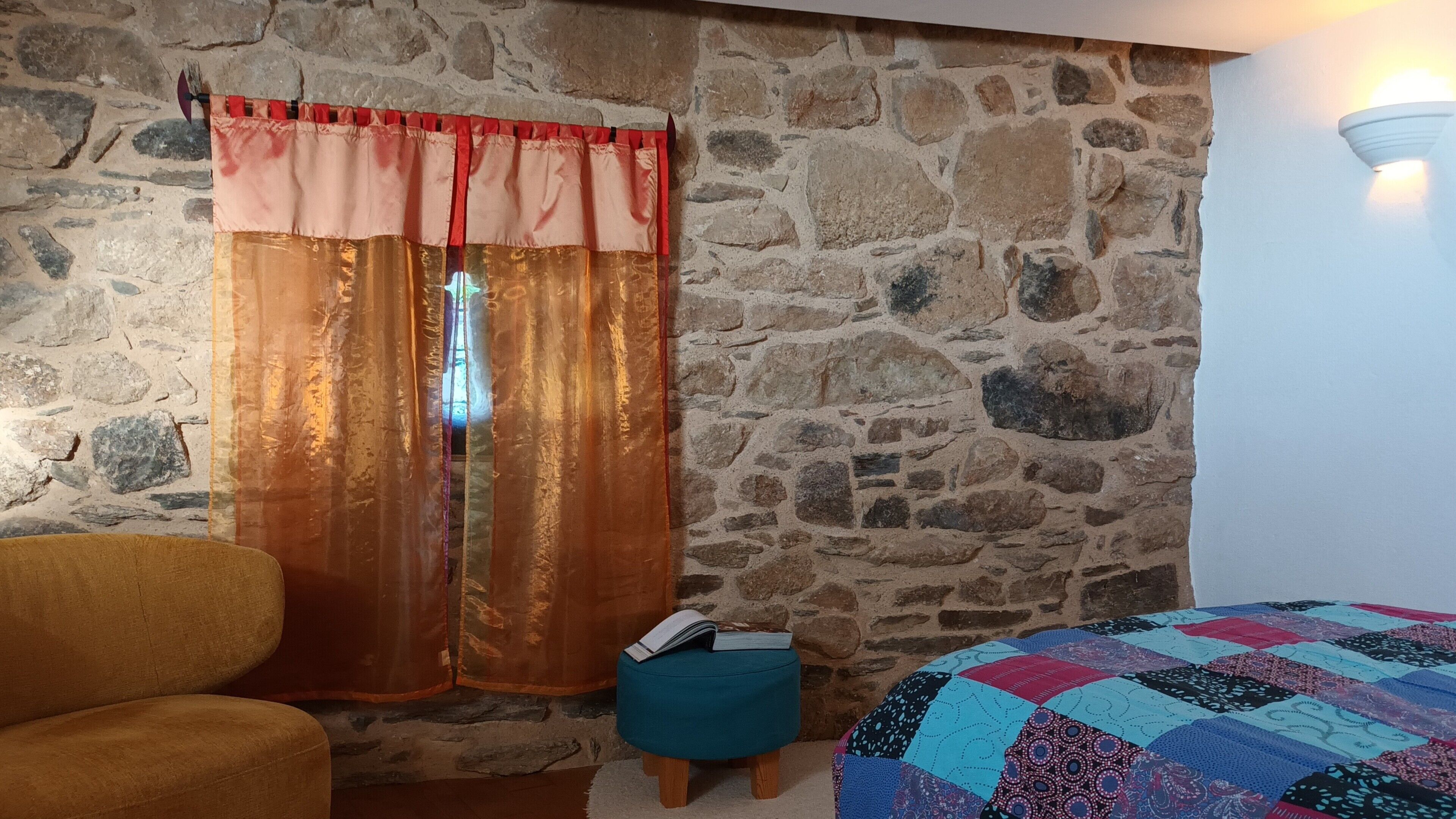 Ein Zimmer mit Steinwand, einem Fenster mit roten und goldenen Vorhängen, einem türkisfarbenen Ottomanen und einem Bett mit einer Patchworkdecke.