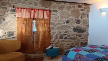A room with a stone wall, a window with red and gold curtains, a teal ottoman, and a bed with a patchwork quilt.