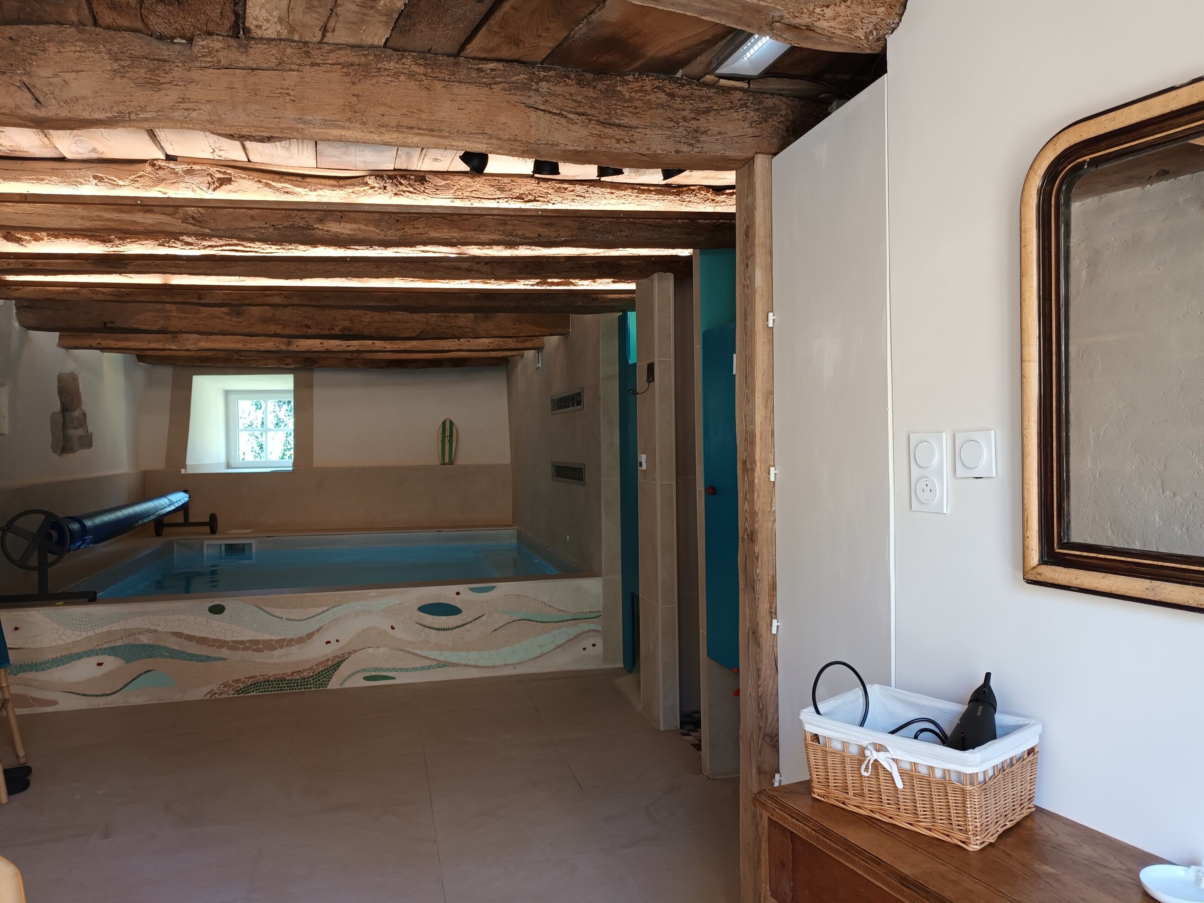 Una piscina interior con techo de madera y suelo de mosaico, una cesta de mimbre sobre una mesa de madera, y un accesorio de iluminación fijado a la pared.