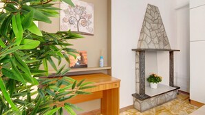 Une pièce avec un bureau en bois, une cheminée en pierre, une plante en pot et un mur orné d’un tableau représentant un arbre.