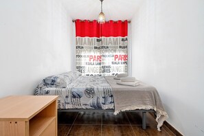Une chambre à coucher avec un lit, une table de chevet en bois, un rideau rouge et une couvre-lit à motifs représentant un paysage urbain.