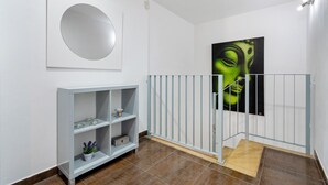 Ein Zimmer mit weißer Wand, einem runden Spiegel, einem grauen Regal mit Dekorationsobjekten, einem grünen abstrakten Gemälde und einem weißen Geländer.