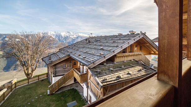 A wooden chalet with a sloped roof, a balcony, and a view of snow-capped mountains.