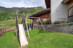 Estructura de juego de madera con un tobogán, un muro de piedra y una valla de madera en un patio cubierto de césped, con una montaña de fondo.