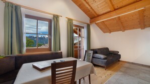 A room with a dining table, a sofa, a window with curtains, and a view of mountains.
