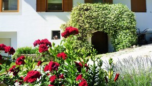 A white building with a covered entrance, red flowers in the foreground, and a garden with various plants. - Farmhouse 'Fröhlichhof Pergola' with Mountain View, Shared Pool and Wi-Fi (Algund)