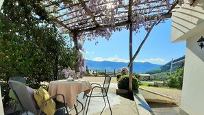 A patio with a table and chairs, a pergola with flowering vines, and a view of mountains. - Farmhouse 'Fröhlichhof Pergola' with Mountain View, Shared Pool and Wi-Fi (Algund)
