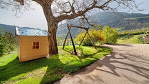 A wooden cabin with a window, a swing set, and a paved path leading to a gate. - Farmhouse 'Fröhlichhof Wohnung Panorama' with Mountain View, Shared Pool and Wi-Fi (Algund)