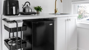 Une cuisine de taille réduite avec des meubles blancs, un réfrigérateur noir, une machine à café et une bouilloire posés sur le plan de travail.