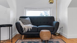 Un salon moderne avec un canapé gris foncé, un petit pouf rond en osier, une table d’appoint noire et un grand lampadaire.