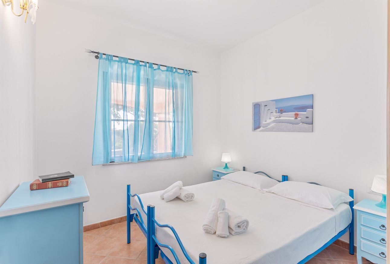 Ein Schlafzimmer mit Bett, Schreibtisch, Stuhl, Fenster mit blauen Vorhängen, Nachttisch und einem Wandbild.