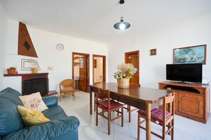 A living room with a sofa, a TV, a dining table with chairs, and a kitchen area in the background.
