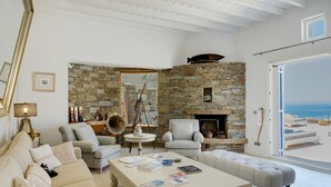 A spacious living room with a stone fireplace, a large window overlooking the sea, and a cozy seating area. - Villa 'Blueros Luxury' with Sea View, Wi-Fi and Air Conditioning (Syros)