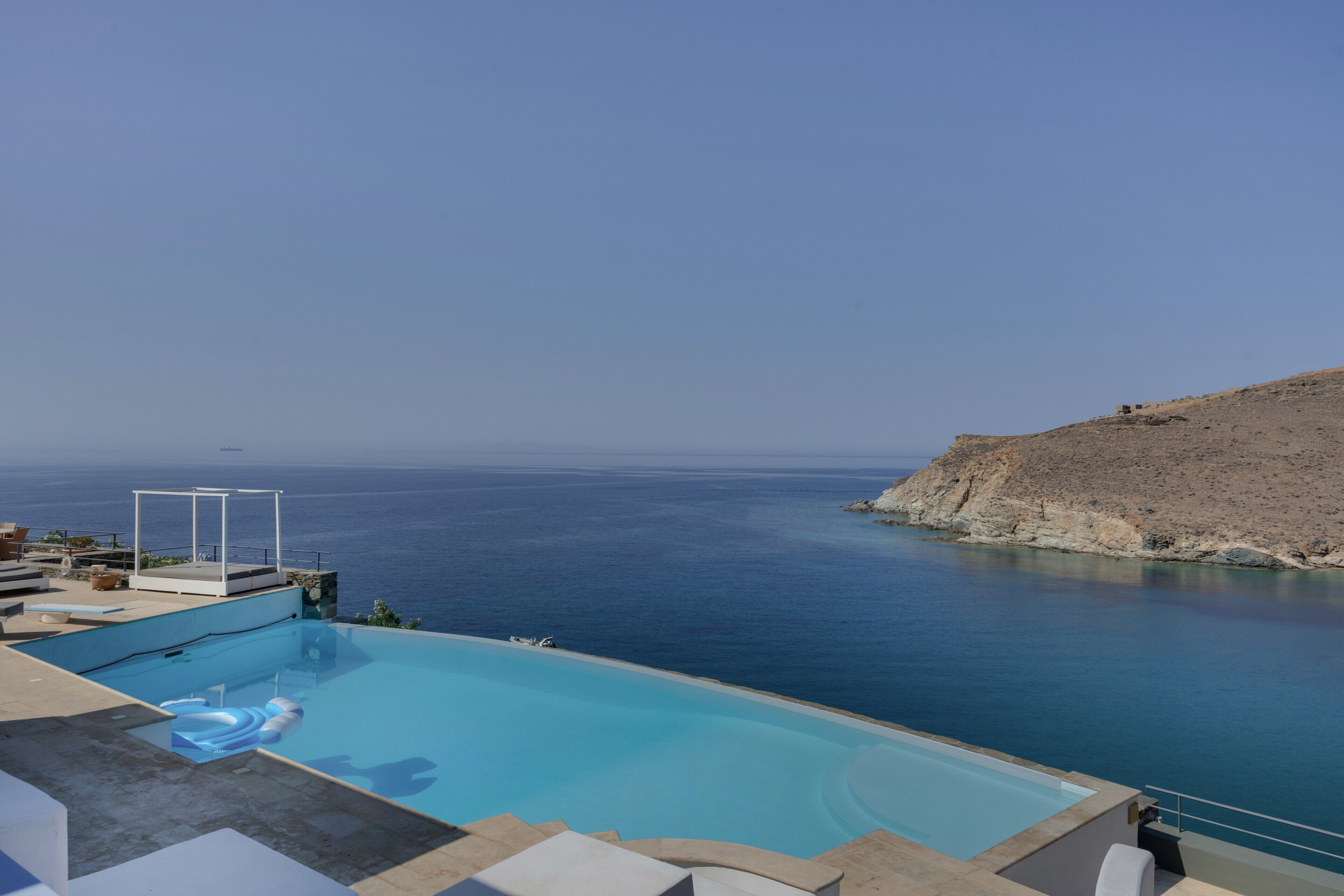 Ein Schwimmbecken mit Blick auf das Meer, ein weißes Pavillon und eine felsige Hügelterrasse.